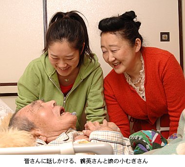 写真：ベッドに寝ている晋さんに、鶴英さんと娘のむぎさんが笑顔で語りかける