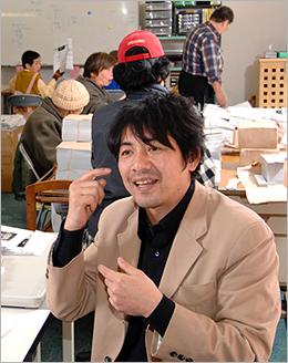 写真：手話で話している岩本さん。後ろでは作業所に通うなかまたちが紙袋の折り曲げの作業をしている