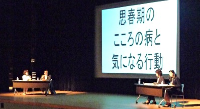 写真：第二部の舞台の様子。真中にスクリーン、左側に司会の河野さん、青木さん、右側に林さん、新開さん