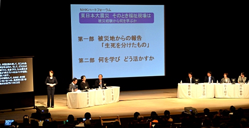 写真：舞台上の様子。右から、内出さん、井上さん、菊池さん、青田さん、立木さん、湯浅さん、町永さんの順で座っている。町永さんの隣には手話通訳者、そしてパソコン要約筆記の画面がある。