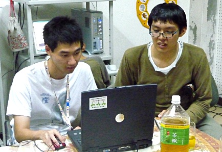 写真：1台のパソコンの前に2人のメンバーが座って、看板作りをしている。