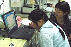 写真：パソコンで看板を製作しているやすのさんと、その様子を見守る小西 栄理さん